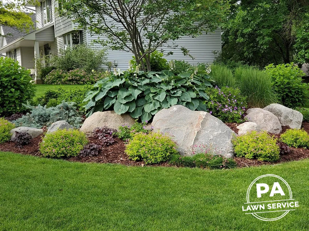 A beautifully designed residential front yard with a landscaped garden bed featuring mulch, rocks, and various plants.